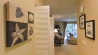 This single-story space offers a warm and inviting atmosphere, highlighted by a coastal-inspired aesthetic that carries throughout the living area. A distinctive white louvered shutter partition serves as a charming architectural detail, subtly dividing the entryway from the main living space while adding character and visual interest to the interior.

The living area is finished with plush wall-to-wall carpeting in a neutral tone, contributing to a comfortable and relaxed ambiance. Warm beige walls provide a soft backdrop that complements the coastal-themed wall art, which features shell and fossil motifs in a mosaic-style arrangement, adding a decorative focal point near the entry.

The space is furnished with a comfortable seating arrangement centered around a dark wood accent table, creating an intimate gathering area. Natural light flows generously into the room through a large sliding glass door dressed with elegant draping curtains, offering views of a lush outdoor area and providing direct access to what appears to be a private patio or deck. The overall design balances casual comfort with thoughtful decorative touches, resulting in a welcoming and cohesive living environment.