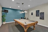 Located at 30300 Antelope Rd in Menifee, CA, this single-floor game room is a well-appointed recreational space designed for entertainment and leisure. The room is anchored by a full-size billiard table with ornate carved legs and a light grey felt surface, positioned beneath a striking multi-shade industrial-style pendant light fixture that provides focused illumination over the playing surface. Recessed ceiling lighting throughout ensures the entire space is evenly and brightly lit.

The room features a two-tone wall design, with a cool slate-blue accent wall contrasting against the surrounding light grey and white walls, lending the space a modern and inviting ambiance. Dark, low-pile carpeting covers the floor, adding warmth and absorbing sound throughout the room. A wall-mounted flat-screen television is positioned in the corner, complemented by decorative domino-style wall art on the adjacent wall.

A compact bistro-style table with two sleek black chairs provides casual seating for guests, while potted plants add touches of greenery. A decorative wooden cue rack and a patriotic-themed canvas print contribute character and personality to the space. A doorway connects to an adjacent room, enhancing the overall functionality of this dedicated recreational area.