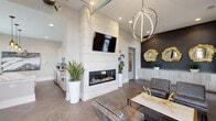 Located at 3350 W Friendly Ave in Greensboro, NC, this single-floor space delivers a refined, contemporary aesthetic designed for both comfort and functionality. The open layout is anchored by a dramatic floor-to-ceiling white tile fireplace with a wall-mounted television above, creating an immediate focal point upon entry. Large-format tile flooring flows seamlessly throughout, unifying the various areas of the space.

The lounge area features high ceilings that amplify the sense of openness, furnished with charcoal leather seating arranged around a marble-topped coffee table with gold accent décor. A striking spherical metallic chandelier serves as a design centerpiece overhead. One wall is clad in dark subway tile and adorned with three ornate gold-framed mirrors, adding depth and visual contrast.

The kitchen area is well-appointed with light wood cabinetry, granite countertops, a gray subway tile backsplash, and stainless steel appliances including a refrigerator and microwave. A large island provides additional prep and gathering space, while pendant lighting adds a warm, modern touch.

Adjacent to the kitchen, a dedicated dining area accommodates a full-sized table with seating for several guests, bathed in natural light from tall, draped windows that frame exterior views. The overall ambiance is modern and sophisticated.
