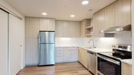 This single-floor unit showcases a clean, contemporary design with an open-concept layout that connects the living area to the kitchen. Light wood-style flooring runs throughout, lending warmth and visual continuity to the space.

The kitchen is well-appointed with flat-front cabinetry in a light wood finish, extending from floor to ceiling to maximize storage. A white subway tile backsplash and light-toned quartz countertops provide a bright, polished aesthetic. Stainless steel appliances include a full-size refrigerator, dishwasher, and range, while open shelving and recessed lighting add functional detail. An under-cabinet range hood completes the cooking area.

The living space is generously proportioned, with large windows and a sliding glass door that invite natural light and offer views of surrounding greenery. The open sightlines between the living area and kitchen create an airy, connected feel.

The bedroom features wood-style flooring, a window with outdoor views, and a closet. The bathroom maintains the unit's clean palette with white walls, wood-style flooring, chrome fixtures, a wall-mounted grab bar, and a blue-accented vanity with a white countertop.