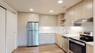 This single-floor unit showcases a clean, contemporary design with an open-concept layout that connects the living area to the kitchen. Light wood-style flooring runs throughout, lending warmth and visual continuity to the space.

The kitchen is well-appointed with flat-front cabinetry in a light wood finish, extending from floor to ceiling to maximize storage. A white subway tile backsplash and light-toned quartz countertops provide a bright, polished aesthetic. Stainless steel appliances include a full-size refrigerator, dishwasher, and range, while open shelving and recessed lighting add functional detail. An under-cabinet range hood completes the cooking area.

The living space is generously proportioned, with large windows and a sliding glass door that invite natural light and offer views of surrounding greenery. The open sightlines between the living area and kitchen create an airy, connected feel.

The bedroom features wood-style flooring, a window with outdoor views, and a closet. The bathroom maintains the unit's clean palette with white walls, wood-style flooring, chrome fixtures, a wall-mounted grab bar, and a blue-accented vanity with a white countertop.