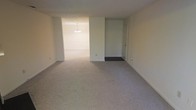 This single-story space presents a clean, open layout with a neutral design aesthetic. The main living area is finished with wall-to-wall carpeting in a light gray-beige tone, providing a soft, uniform foundation throughout. Smooth, light-colored walls complement the carpeting, creating a bright and airy atmosphere even without abundant natural light.

An open passageway connects the main living area to an adjoining room, which features an elegant chandelier as a focal point, suggesting use as a dining area. The chandelier adds a subtle decorative element to an otherwise understated space. The transition between rooms is defined by a subtle step or threshold, differentiating the two areas while maintaining an overall sense of openness.

A doorway on the right side of the main room, fitted with a white door, leads to additional areas of the unit. The consistent use of white trim, neutral wall tones, and carpeted flooring throughout creates a cohesive, move-in-ready environment. The straightforward layout and simple finishes make this space highly adaptable to a variety of furniture arrangements and interior design styles.