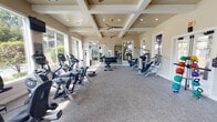 Located at 3601 Grapevine Mills Pkwy Frontage Rd in Grapevine, TX, this well-appointed fitness center offers a comprehensive workout environment designed for both cardio and strength training. The single-floor facility features a distinctive coffered ceiling with recessed lighting and ceiling fans, contributing to a bright, airy atmosphere throughout the space. Large floor-to-ceiling windows line the walls, flooding the interior with abundant natural light and providing views of the surrounding outdoor amenities, including a pool area and landscaped grounds. The main workout area is equipped with a full lineup of cardio machines, including stationary bikes, ellipticals, and treadmills, all positioned to take advantage of the outdoor views. A dedicated strength training section houses a multi-station cable machine, a weight bench, and a full rack of dumbbells in varying sizes. Full-length mirrors expand the sense of space and allow for form checks during exercise. A separate area features an assortment of medicine balls and a suspension training strap, catering to functional fitness routines. A dual-unit water fountain station is conveniently located near the exit for easy hydration. The neutral-toned walls, carpeted flooring, and clean, well-maintained equipment combine to create an inviting and functional fitness environment.