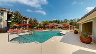 This outdoor amenity space in Daphne, Alabama, centers on a generously sized resort-style swimming pool with clear, blue-green water and a smooth concrete surround. The pool area is well-appointed with numerous lounge chairs arranged along the deck, offering ample space for sunbathing beneath the open Alabama sky. Red-accented pergolas and covered cabanas are positioned throughout the deck, providing shaded retreats and contributing a cohesive, vibrant aesthetic to the space. A separate spa or water feature is visible adjacent to the main pool, adding to the recreational appeal. The surrounding landscape includes mature trees and palm plantings, and a wrought iron perimeter fence encloses the area for security and privacy. Multistory residential buildings are visible in the background, framing the space.

Adjacent to the pool deck is a single-story covered utility and amenity structure. Its exterior features board-and-batten siding in a muted sage tone, accented by a brick base and a teal six-panel door. Inside, the covered area houses a vending machine and provides a practical service point for pool visitors. The overall atmosphere of the space is open, well-maintained, and designed for outdoor leisure and community enjoyment.