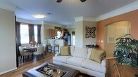 This single-story residence showcases an open-concept layout that integrates the living room, dining area, and kitchen into one cohesive, flowing space. The living room is anchored by a plush white sofa accented with gold and green throw pillows, complemented by a leather ottoman that doubles as a coffee table. Crown molding runs along the ceiling, adding an elegant architectural detail, while a ceiling fan overhead ensures comfort. The accent wall in a warm terracotta tone adds a bold design element, and decorative wall art enhances the overall aesthetic.

The kitchen is visible from the living area and features wooden cabinetry, a stainless steel refrigerator, and a raised breakfast bar with tall seating — ideal for casual dining or entertaining. Pendant lighting hangs above the bar, adding a stylish focal point. Adjacent to the kitchen, the dining area accommodates a full table and chair set, positioned near draped windows that allow natural light to fill the space. Hardwood-style flooring flows consistently throughout, tying each zone together and contributing to a warm, welcoming ambiance.