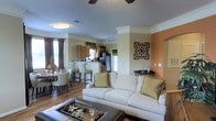 This single-story residence showcases an open-concept layout that integrates the living room, dining area, and kitchen into one cohesive, flowing space. The living room is anchored by a plush white sofa accented with gold and green throw pillows, complemented by a leather ottoman that doubles as a coffee table. Crown molding runs along the ceiling, adding an elegant architectural detail, while a ceiling fan overhead ensures comfort. The accent wall in a warm terracotta tone adds a bold design element, and decorative wall art enhances the overall aesthetic.

The kitchen is visible from the living area and features wooden cabinetry, a stainless steel refrigerator, and a raised breakfast bar with tall seating — ideal for casual dining or entertaining. Pendant lighting hangs above the bar, adding a stylish focal point. Adjacent to the kitchen, the dining area accommodates a full table and chair set, positioned near draped windows that allow natural light to fill the space. Hardwood-style flooring flows consistently throughout, tying each zone together and contributing to a warm, welcoming ambiance.