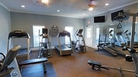 This fitness center offers a dedicated space for both cardio and strength training workouts. The room is finished with neutral grey walls, crown molding, and a textured ceiling, giving it a clean and functional aesthetic. Recessed lighting and a ceiling fan with a light fixture ensure the space remains well-lit and comfortable during exercise sessions. Multiple windows along the walls allow natural light to fill the room, creating an open and energizing atmosphere. The cardio section is outfitted with a treadmill, an elliptical machine, and additional upright exercise equipment, all positioned to allow ample space for use. A wall-mounted flat-screen television is positioned for easy viewing during workouts. A large mirror along one wall visually expands the space and aids in monitoring form during exercise. Strength training benches are available for free weight exercises. Informational signage, including fitness center rules, is posted on the wall for member reference. The flooring includes protective matting beneath the equipment, promoting safety and equipment stability. The space is accessed through a dedicated entryway door, reinforcing its role as a self-contained amenity facility.