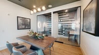 Located at 5555 W Ina Rd in Marana, Arizona, this contemporary dining room is a thoughtfully designed, single-floor space that blends modern aesthetics with functional versatility. The room centers around a striking live-edge wood dining table paired with stylish teal and gray upholstered chairs, creating a warm yet sophisticated gathering area. A linear pendant light fixture with globe bulbs hangs overhead against a deep olive-green ceiling, adding a bold design contrast to the crisp white walls below. Light wood-look flooring runs throughout, lending warmth and continuity to the space. Abstract framed artwork adorns the walls, contributing to the curated, upscale atmosphere. A defining architectural feature is the floor-to-ceiling glass partitions with bold black geometric patterned decals, which separate the dining area from adjacent private rooms. These glass-walled rooms offer visual transparency while maintaining acoustic separation, each accessed via sleek matte black door handles. The adjoining lobby area, partially visible through the glass, features soaring ceilings, large windows with natural light, and desert-inspired plantings, connecting the interior to the Arizona landscape beyond. The overall ambiance is polished, modern, and welcoming — ideal for meetings, private dining, or community gatherings.