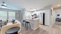 This thoughtfully designed single-floor residence offers two bedrooms, one bathroom, and a seamlessly connected living and kitchen space. Light-toned walls, wood-look flooring, and abundant natural light from multiple windows create a bright, airy atmosphere throughout.

The open-concept kitchen is equipped with white shaker cabinetry, stainless steel appliances including a refrigerator, range, and microwave, and a white countertop peninsula with bar seating — ideal for casual dining or entertaining. A skylight adds additional natural brightness to the kitchen area.

The living room flows naturally from the kitchen, providing a comfortable gathering space with a ceiling fan and room for a full seating arrangement. An entry foyer with a console table creates a welcoming transition into the home.

Both bedrooms feature soft carpet flooring, ceiling fans, and windows with natural views. The primary bedroom offers ample space for a full bedroom suite along with a dedicated workspace area.

The bathroom features white shaker-style cabinetry, a white countertop vanity with an undermount sink, brushed nickel fixtures, and a large mirror. A dedicated closet provides convenient storage. Clean lines and a neutral palette unify every room, resulting in a cohesive and functional living environment.