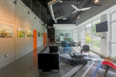 Located at 1785 Almaden Road in San Jose, CA, this single-floor commercial space offers a compelling blend of industrial aesthetics and modern functionality. The open-plan layout is anchored by soaring ceilings with fully exposed ductwork, structural elements, and dark-painted overhead infrastructure, lending a distinctly urban, loft-like character throughout. Large floor-to-ceiling windows and glass storefront walls line the exterior, flooding the interior with natural light and providing unobstructed views of the surrounding greenery.

The polished concrete floors run continuously through the space, reinforcing the industrial design language while remaining visually clean and refined. A lounge area near the entrance features tufted leather sofas, a low-profile coffee table, and colorful accent chairs, creating an inviting gathering spot beneath large ceiling fans. Art pieces displayed along the textured concrete walls add a gallery-like quality to the environment.

A glass-enclosed private office or meeting room provides visual transparency while offering acoustic separation. Adjacent to it, a conference area with a sleek table and chrome-framed chairs sits directly beside the window wall, bathed in daylight. Vibrant orange accent doors punctuate the neutral palette, adding bold contrast. A utility area with storage cabinetry and equipment completes the space. Overall, the design balances creative energy with practical workspace versatility.