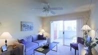 This single-story space centers around a comfortable living room designed for both relaxation and functionality. The room features carpeted flooring and warm, neutral-toned walls that create a welcoming ambiance. A ceiling fan with an integrated light fixture is mounted overhead, providing both air circulation and supplemental lighting throughout the space.

The living area is furnished with a plush blue-gray sofa accented with decorative purple pillows, paired with a sleek, minimalist black coffee table that anchors the seating arrangement. A small side table with a table lamp adds a warm, ambient glow to the corner of the room, complementing the overall inviting atmosphere.

A standout feature of this space is the sliding glass door, which floods the living room with natural light and provides direct access to an outdoor patio area. Through the glass, greenery and an open-air balcony railing are visible, offering a pleasant outdoor connection. The patio door is fitted with a welcome mat, suggesting a seamless transition between interior and exterior living. Wall art depicting a city skyline at night adds a decorative focal point, contributing to the space's modern, casual character.