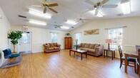 Located at Fountainhead Circle in Melbourne, Florida, this single-floor community space serves as both a leasing office and a resident gathering area. The entrance greets visitors with clean white trim, crown molding, hardwood-style flooring, and a welcoming mat that sets an organized, professional tone. A leasing office is clearly marked and accessible directly from the entryway, providing a functional hub for resident services.

The spacious living room is the heart of the space, featuring soaring vaulted ceilings that amplify the sense of openness throughout. Multiple ceiling fans are evenly distributed across the ceiling, ensuring comfortable airflow. Warm hardwood-style flooring spans the entire area, lending a cohesive and inviting aesthetic. Natural light filters in through several windows fitted with vertical blinds, brightening the cream-toned walls accented by a classic chair rail detail.

The living room is furnished with comfortable upholstered sofas and a loveseat arranged around a coffee table, creating a relaxed lounge setting. A wooden armoire, decorative plants, and accent artwork add warmth and character to the room. Additional seating and table space along one side of the room offer flexibility for various uses. The open floor plan allows for easy movement throughout the space, making it well-suited for community gatherings and resident interactions.