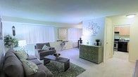 This single-floor space offers a comfortable and thoughtfully arranged living environment defined by a warm, neutral color palette and an open, airy atmosphere. The main living area is generously proportioned, featuring plush wall-to-wall carpeting that adds a soft, welcoming feel underfoot. Large windows dressed with vertical blinds line the walls, allowing abundant natural light to fill the room while providing privacy and light control as needed. The living room is anchored by a spacious seating arrangement, complemented by a dark-framed coffee table and a patterned area rug that adds texture and visual interest to the space. A decorative green sideboard, adorned with tasteful accents and greenery, serves as a standout furniture piece along one wall, paired with an ornate metal wall art installation that lends a touch of artistic character to the room. A secondary seating area with a small accent table is positioned near the windows, creating a versatile nook ideal for reading or casual use. The overall ambiance is calm and inviting, with soft tones throughout the walls and ceilings contributing to a light, open feel. The layout flows naturally, making the space functional and easy to navigate across its single level.