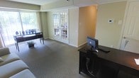 This single-floor space presents a functional and professionally appointed interior designed to accommodate workstation use. The room features neutral wall tones, combining soft green and warm beige, which contribute to a calm, focused atmosphere. Carpeted flooring in a muted gray-brown shade runs throughout, adding comfort underfoot and helping to define the space as a quiet, professional environment.

A standout architectural feature is the set of white-framed French doors positioned centrally along one wall, offering views to an exterior area and allowing natural light to filter into the room. These doors provide both a visual connection to the outdoors and a sense of openness within the otherwise enclosed space.

Along one side, a dark wood desk supports a desktop computer and keyboard, serving as a dedicated workstation. Near the window, an additional dark console table provides supplementary workspace or display surface. A security camera is mounted near the ceiling, indicating this is a monitored, shared-use environment.

Overall, the space functions well as a business center or computer lounge, offering a tidy, organized setting with adequate lighting and a straightforward, welcoming layout.