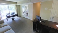 This single-floor space presents a functional and professionally appointed interior designed to accommodate workstation use. The room features neutral wall tones, combining soft green and warm beige, which contribute to a calm, focused atmosphere. Carpeted flooring in a muted gray-brown shade runs throughout, adding comfort underfoot and helping to define the space as a quiet, professional environment.

A standout architectural feature is the set of white-framed French doors positioned centrally along one wall, offering views to an exterior area and allowing natural light to filter into the room. These doors provide both a visual connection to the outdoors and a sense of openness within the otherwise enclosed space.

Along one side, a dark wood desk supports a desktop computer and keyboard, serving as a dedicated workstation. Near the window, an additional dark console table provides supplementary workspace or display surface. A security camera is mounted near the ceiling, indicating this is a monitored, shared-use environment.

Overall, the space functions well as a business center or computer lounge, offering a tidy, organized setting with adequate lighting and a straightforward, welcoming layout.