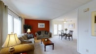 This single-floor space features an open-concept design that seamlessly connects the living and dining areas into one cohesive, functional layout. The living room is anchored by a striking burnt-red accent wall, which adds warmth and visual contrast against the surrounding neutral white walls and light-toned carpeting. Comfortable upholstered seating is arranged around a classic wooden coffee table, creating an inviting area for relaxation. Framed artwork and a decorative floor lamp contribute to the room's lived-in, welcoming ambiance. Crown molding along the ceiling adds a refined architectural detail throughout the space. The dining area sits adjacent to the living room and is furnished with a glass-top table and wrought-iron style chairs, offering an elegant yet understated dining setting. A ceiling fan with a light fixture overhead provides both air circulation and ambient lighting. French doors at the rear of the dining area allow natural light to fill the space and provide access to an outdoor area, enhancing the overall sense of openness. The consistent carpeting and unified color palette throughout tie both areas together harmoniously.