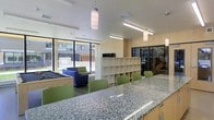 This single-floor communal space is designed to serve as a multifunctional gathering and activity hub. At the forefront, a generously sized granite countertop bar anchors the room, complemented by a row of green chairs that invite casual seating and socializing. Overhead, a combination of pendant lights and linear fluorescent fixtures provides ample, well-distributed illumination throughout the space.

Moving deeper into the room, a set of light wood open shelving units offers organized storage and display areas, while an adjacent dark metal shelving unit adds a contrasting design element. A television is mounted atop the shelving structure, further enhancing the space's utility as an entertainment and study area.

To one side, a lounge zone features a pool table and comfortable blue upholstered chairs, creating a relaxed recreational corner. Large floor-to-ceiling windows and glass doors line the perimeter, flooding the interior with natural light and offering clear views of a landscaped outdoor courtyard and surrounding building facades. The clean white ceilings, neutral tones, and light wood finishes throughout give the space a bright, modern, and welcoming ambiance suited for a variety of communal activities.