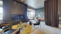 Located at 2626 Throckmorton Street in Dallas, TX, this 990-square-foot amenity space at Griffis Residential is a thoughtfully designed community lounge spanning a single floor. The entrance greets visitors with a dramatic corridor featuring polished concrete floors, deep charcoal accent walls, and steel-framed glass doors that set an upscale, contemporary tone.

The main lounge area is anchored by a sleek linear fireplace set into dark tile walls, complemented by mid-century modern leather armchairs, a geometric glass coffee table, and warm area rugs. A striking hand-painted botanical mural in muted blue-gray tones adorns one wall, adding an artistic focal point alongside a cognac leather sofa. Steel-framed glass partitions allow natural light to flow throughout the space.

The kitchen and dining area features floor-to-ceiling dark marble-look tile, white upper cabinetry with under-cabinet lighting, a matte black countertop with a brass-accented faucet, stainless steel appliances, and a large communal dining table beneath a sculptural looping pendant light.

The outdoor terrace offers a covered seating area with a living green wall displaying a neon "It's about you" sign, creating a welcoming, resort-style atmosphere ideal for relaxation and community gatherings.