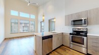 This single-floor residence showcases a contemporary design defined by dramatically high ceilings that create an expansive, airy atmosphere throughout the living areas. The open-concept layout connects the living room to the kitchen seamlessly, with warm hardwood-style flooring running throughout the main living spaces and a door providing direct access to an outdoor area.

The kitchen is well-appointed with light wood cabinetry extending to the elevated ceilings, stainless steel appliances including a refrigerator, range, and dishwasher, and a peninsula with a white quartz countertop and undermount sink. Pendant lighting adds a refined touch overhead. A ceiling fan serves the living area, which benefits from oversized windows drawing in generous natural light and views of the surrounding neighborhood.

The bedrooms feature plush carpeting and large windows with outdoor views, with one room benefiting from soaring ceiling height and clerestory windows. The bathroom includes a light wood vanity, white quartz countertop, and a full tub/shower combination. A dedicated laundry room accommodates a full-size side-by-side washer and dryer with overhead shelving for additional storage.