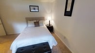 This single-floor bedroom is designed with comfort and simplicity in mind. The room features neutral, light beige walls that contribute to a serene and restful ambiance. Soft wall-to-wall carpeting in a warm tan hue adds a cozy feel underfoot and complements the overall color palette.

The space is furnished with a queen-sized bed dressed in crisp white linens and accented with decorative pillows, positioned against an upholstered headboard in a subtle gray tone. A dark-finished nightstand sits beside the bed, topped with a warm-glowing table lamp that provides soft, intimate lighting. At the foot of the bed, a tufted dark leather bench adds a functional and stylish touch.

Two framed artworks adorn the walls — one positioned above the bed and another on the adjacent wall — adding visual interest without overwhelming the minimalist aesthetic. The wide baseboards provide clean architectural definition throughout the room. The overall design prioritizes a calm, uncluttered environment, making it a quiet and inviting retreat well-suited for rest and relaxation.