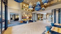This expansive single-floor amenity space is defined by its dramatic double-height ceilings and an eye-catching array of oversized blue woven pendant lights that serve as the central focal point of the main lobby. The light-toned herringbone hardwood flooring anchors the space with a refined yet approachable aesthetic, while floor-to-ceiling black-framed glass windows and doors flood the interior with natural light and provide direct views of the surrounding outdoor courtyard.

The main lounge area features curated built-in display shelving with warm wood tones, showcasing decorative art objects and sculptures. Seating vignettes are thoughtfully arranged throughout, incorporating navy blue sofas with mustard yellow accents, leather sling chairs, and ottomans. A secondary lounge area transitions into a more intimate setting with a stone accent wall, upholstered armchairs, and a sculptural arc floor lamp.

A dedicated social lounge offers a fireplace, mixed seating arrangements, and art-adorned walls. Adjacent to this is a well-appointed kitchenette featuring panel-ready appliances, open display cabinetry, a circular bar-height island with seating, and pendant lighting overhead. Throughout, the design balances contemporary sophistication with warm, textural materials.