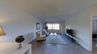 This single-floor space offers a clean, modern aesthetic with a thoughtfully designed open layout. The living area serves as the centerpiece, featuring smooth hardwood flooring that extends throughout, lending warmth and continuity to the space. Crisp white walls and a neutral color palette create a bright, airy atmosphere that feels welcoming and uncluttered.

A large picture window dominates one wall of the living area, flooding the room with natural light and offering a pleasant view of lush, green foliage outside — a refreshing connection to the outdoors. The living space is furnished with a classic white armchair and a matching sofa, centered around a sleek glass coffee table and anchored by a plush area rug, creating a comfortable and cohesive seating arrangement.

On the opposite side of the room, a large flat-screen television is mounted above a low-profile media console, adding a functional entertainment focal point. A decorative indoor plant adds a touch of organic texture near the window, complementing the natural tones of the space. Tasteful artwork adorns the walls, contributing to the refined, contemporary character of the interior. Overall, the space balances simplicity and comfort with a polished, livable design.