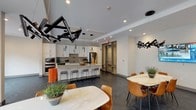 The club room at 150 Glover Ave in Norwalk, CT is a thoughtfully designed communal space that blends functionality with contemporary style. The fully equipped kitchen anchors one end of the space, featuring white shaker cabinetry, granite countertops, stainless steel appliances including a full-size refrigerator and dishwasher, and dark herringbone-patterned tile flooring. A large granite-topped island with bar seating serves as a central gathering point, accented by striking sculptural pendant lighting. Multiple dining areas are arranged nearby, each with its own distinct table and seating configuration, creating flexible options for both casual and social settings. The lounge area transitions seamlessly into the space, offering a generous sectional sofa on patterned carpeting, wall-mounted flat-screen TV, and floor-to-ceiling windows with sliding glass doors that open to an outdoor patio. A bold teal accent wall adds a vibrant focal point to this area. Beyond the lounge, a game room features a pool table, a world map wall mural, and additional seating, contributing an entertaining dimension to the space. Throughout, recessed lighting, modern fixtures, and clean architectural lines give the club room a polished, contemporary ambiance ideal for community gatherings.