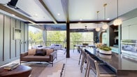 This thoughtfully designed single-floor space seamlessly blends indoor comfort with outdoor living, creating an ideal environment for entertaining and relaxation. The layout flows effortlessly from the interior living area to a generously sized covered terrace, unified by consistent tile flooring and exposed dark wood beam ceilings that lend warmth and architectural character throughout.

The indoor lounge area features sage-green board-and-batten wall paneling, a wall-mounted flat-screen television, and a ceiling fan, complemented by an elegant arched architectural detail that frames a decorative accent wall. Recessed lighting and pendant fixtures with brass accents provide layered illumination throughout the space.

The compact kitchenette is outfitted with sage-green cabinetry, dark stone countertops, stainless steel appliances including a wall oven, microwave, and French-door refrigerator, and a striking ribbed tile backsplash that adds texture and visual depth. A long bar-height counter with woven rattan seating offers a casual dining option.

The expansive covered terrace is the centerpiece of the space, featuring overhead infrared heaters, a ceiling fan, and open railings that frame panoramic views of mature tree canopies. Multiple seating arrangements and a dining table make it well-suited for both intimate gatherings and larger events, with the lush greenery providing a serene natural backdrop year-round.