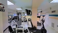 This fitness center offers a comprehensive workout environment spread across a single level. The space is outfitted with a diverse selection of exercise equipment, including treadmills, stationary bikes, ellipticals, and multi-use cable strength machines, catering to both cardio and resistance training needs. The room features a clean white color scheme accented by a teal horizontal stripe along the walls, giving the space a crisp, energetic atmosphere. Recessed panel lighting in the ceiling ensures even illumination throughout, while small windows along one wall allow for natural light to supplement the interior lighting. A wall-mounted flat-screen television provides entertainment during workouts. Structural columns are integrated into the layout, subtly dividing the space into distinct equipment zones without interrupting the overall open feel. The flooring is smooth and light-toned, contributing to the bright and airy ambiance. The facility appears well-maintained and organized, making it a practical and inviting space for residents or guests seeking a convenient on-site fitness option.