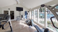 This single-story fitness center offers a thoughtfully designed workout environment that balances functionality with an open, inviting atmosphere. The space features vaulted ceilings that lend a sense of airiness, while large multi-pane windows along one wall flood the interior with natural light and provide views of the surrounding outdoor area, including glimpses of a pool and landscaping.

The flooring consists of a herringbone-patterned wood-look surface, adding a polished aesthetic throughout. Full-length mirrors line portions of the walls, visually expanding the space and supporting proper form during exercise. A ceiling fan and wall-mounted flat-screen televisions enhance comfort during workouts.

Cardio equipment is well-represented, with multiple treadmills, elliptical trainers, and a stationary bike arranged throughout the room. A multi-station cable and strength machine occupies a dedicated area on protective rubber matting, while a dumbbell rack provides free weight options. Motivational wall decals, including "Be Active, Be Healthy, Be Happy" and "The Body Achieves What the Mind Believes," contribute to an encouraging atmosphere. The overall design is clean, bright, and purpose-driven.