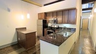 This single-floor space centers around a well-appointed kitchen that combines functionality with modern design. The kitchen features dark granite countertops that extend across both the main counter and a central island, providing generous workspace for meal preparation. Rich, warm-toned wood cabinetry lines the walls, offering substantial storage through both upper and lower cabinets. Stainless steel appliances, including a refrigerator, electric range, and built-in microwave, lend a contemporary feel to the space. A stainless steel sink is integrated into the island, adding convenience to the layout. Pendant lighting hangs above the island, contributing soft, focused illumination that complements the overall aesthetic. The flooring appears to be polished concrete, a detail that adds an industrial character to the space. The high ceilings, visible in the background, enhance the sense of openness and volume throughout. The neutral wall tones allow the darker cabinetry and countertops to stand out as focal points, creating a balanced and cohesive atmosphere throughout the kitchen area.