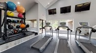 This fitness center is designed to support a full range of workout routines within a bright, open environment. The space is defined by its striking vaulted ceiling, which rises dramatically overhead and is fitted with recessed lighting that evenly illuminates the rubber-floored interior. Large windows span multiple walls, inviting natural light throughout the facility and offering views of the surrounding greenery, creating an energizing atmosphere.

The cardio zone is well-appointed with multiple treadmills, elliptical trainers, and stationary spin bikes, all positioned to take advantage of the outdoor views. Wall-mounted flat-screen televisions are installed near the cardio equipment, providing entertainment during workouts. A dedicated strength-training area features a cable resistance machine, weight benches, and a medicine ball rack stocked with a variety of weighted balls. Full-length mirrors line portions of the walls, enhancing the sense of space and aiding in form correction during exercise.

A structural column anchors the center of the room, subtly delineating the cardio and strength zones. A vending machine is conveniently located near the entrance. The neutral gray and white color palette throughout contributes to the clean, modern aesthetic of this well-rounded fitness facility.