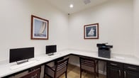 This single-floor business center offers a functional and neatly appointed workspace designed to meet everyday productivity needs. The room is organized around an L-shaped white countertop workstation that wraps along two walls, providing ample desk space for multiple users simultaneously. Several computer monitors with keyboards and mice are positioned along the surface, accompanied by a multifunction laser printer conveniently placed at one end of the counter. Dark wood ladder-back chairs with upholstered seats offer comfortable seating at each workstation.

The walls are finished in a clean, neutral white, lending the room a bright and professional atmosphere, while recessed lighting provides consistent illumination throughout. A patterned carpet in gold and neutral tones adds a subtle decorative touch underfoot. Framed nautical prints depicting sailing vessels are displayed on the walls, contributing a refined, coastal-inspired aesthetic to the space.

A standout architectural feature is the full-height black steel-framed glass partition and door that serves as the room's entryway, offering a modern, sophisticated contrast to the otherwise understated interior. Through the glass, an adjacent common area with striped wallcovering, light wood flooring, and pendant lighting is visible, connecting the business center to the broader space beyond.