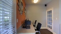 This single-story space functions as a dedicated business center or computer lounge, designed to support focused work in a compact yet functional setting. The room features a built-in counter-height workstation that runs along one wall, providing ample desk space and housing desktop computer equipment along with a keyboard and peripheral accessories. Two ergonomic mesh-back office chairs are positioned at the workstation, accommodating multiple users simultaneously.

The interior design combines a warm terracotta-orange accent wall with neutral gray walls, creating a visually dynamic contrast that adds character to the space. Two decorative sunburst mirrors adorning the orange wall serve as standout artistic elements, lending the room a stylish, curated feel. Overhead, a flush-mount ceiling light fixture provides consistent ambient illumination throughout the space.

Dark carpeting grounds the room and complements the overall color scheme. A small wall-mounted speaker is also present, adding an audio component to the environment. White-framed doors on either side suggest easy access to adjoining areas, contributing to the space's accessibility and connectivity within the broader floor plan.