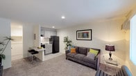 This single-floor unit offers a well-organized layout that efficiently combines living, dining, and kitchen functions within an open-concept design. The living area features neutral carpet flooring, recessed lighting, and light-toned walls that create a bright, welcoming atmosphere. A wall-mounted air conditioning unit provides climate control, while a window along the exterior wall allows natural light and outdoor views into the space.

The kitchen is thoughtfully designed with gray shaker-style cabinetry, granite-look countertops, and black appliances, including a full-size refrigerator and gas range. A peninsula with bar-height seating serves as both a functional prep surface and a casual dining area. Wood-look flooring in the kitchen provides a clean contrast to the carpeted living space.

A hallway connects the main living area to the bathroom and an additional room. The bathroom features a gray vanity with a white countertop, a wall-mounted vanity light, a framed mirror, and a tub-shower combination with gray tile surround. A closet with shelving is positioned adjacent to the bathroom, offering dedicated storage. Overall, the unit maintains a clean, contemporary aesthetic with practical finishes throughout.