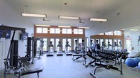 This fitness center is designed to provide a functional and comfortable workout environment. The space is anchored by a generous open floor plan that accommodates a wide variety of exercise equipment, including a row of treadmills and elliptical machines positioned along the perimeter, a dumbbell rack with a full weight selection, and additional strength-training stations. The layout allows for easy movement between equipment without feeling congested.

Architecturally, the room features high ceilings with suspended fluorescent lighting panels that ensure even illumination throughout the space. Ceiling fans are installed to promote air circulation during workouts. A standout feature is the wall of large glass doors and clerestory windows framed in warm orange accents, which flood the interior with natural light and offer views of an outdoor area beyond. The full-length mirror along one wall enhances the sense of space while allowing users to monitor their form. The flooring is a durable, light-toned surface suited for both cardio and weight training. Overall, the space strikes a balance between practicality and an open, energizing atmosphere.