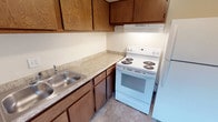 This single-floor unit offers a practical and well-organized layout, with rooms connected by a central hallway that provides easy access throughout the space. The kitchen is efficiently designed, featuring warm wood-toned cabinetry both above and below the counter, light laminate countertops, a stainless steel sink, a white electric coil range with an overhead exhaust hood, and a full-size refrigerator. Light tile flooring completes the kitchen's clean, functional aesthetic. The bathroom includes a wide vanity with a white countertop and an integrated sink, a wood-framed medicine cabinet with mirrored doors, and light flooring that transitions into the carpeted hallway beyond. The living area is generously proportioned, featuring neutral carpet and a ceiling light fixture, with a hallway leading to the bedroom wing. Multiple bedrooms each offer plush carpeting, neutral painted walls, and windows with horizontal blinds that invite natural light into the rooms. The overall ambiance of the unit is clean and move-in ready, with a straightforward, functional design that provides comfortable, practical living across its single level.