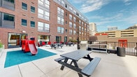 Located at 260 Portland Ave in the heart of downtown Minneapolis, this outdoor courtyard terrace offers a well-appointed communal space set against the backdrop of a classic red brick multi-story building with large grid-pane windows. The terrace is designed to accommodate a variety of activities, making it a versatile amenity for residents and visitors alike.

At one end of the courtyard, a dedicated children's play area features a vibrant red fire truck-themed play structure complete with a slide, set on impact-absorbing rubber surfacing in teal. This family-friendly zone is positioned to allow easy visibility from the surrounding seating areas.

The main terrace area is furnished with multiple dark gray picnic tables paired with benches, offering ample space for outdoor dining and gatherings. Bright red Adirondack-style lounge chairs are arranged in casual clusters, providing comfortable spots for relaxation. A stainless steel grilling station is conveniently situated near the perimeter railing, making outdoor cooking accessible.

Raised planters with ornamental grasses add greenery and a sense of natural warmth to the hardscaped surroundings. The terrace is enclosed by a metal railing system that frames open views of the Minneapolis skyline, including neighboring high-rise towers. Interior common spaces are accessible directly from the courtyard through large glass doors along the brick facade.