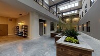 This enclosed atrium is a striking architectural centerpiece defined by its impressive vertical scale, with soaring ceilings that draw the eye upward toward a skylight-equipped roofline, flooding the space with natural light throughout the day. The polished concrete floors extend across the entire area, providing a sleek, industrial-modern foundation that contrasts beautifully with the warmer organic elements found throughout.

Central to the atrium's design are several raised garden planters constructed with dark wood bases and stone-capped edges, filled with lush bamboo, tropical foliage, and ornamental plants. These planters serve as natural dividers, adding a verdant, tranquil quality to the shared space. Painted white brick columns line the perimeter, lending a classic yet contemporary texture that complements the neutral gray wall tones.

Unit entry doors are positioned along the atrium's walls, each accompanied by wall-mounted sconce lighting and black-framed windows that allow residents to connect visually with the courtyard. An upper-level mezzanine with black metal railings overlooks the atrium below. A lounge area near the staircase provides comfortable seating, completing this well-appointed shared environment.