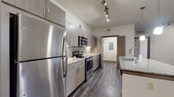 This well-appointed single-floor residence offers a thoughtfully designed layout with modern finishes throughout. The kitchen is a standout feature, showcasing sleek taupe cabinetry, granite countertops, and a full suite of stainless steel appliances including a refrigerator, electric range, and built-in microwave. A mosaic tile backsplash adds a stylish accent, while track lighting and pendant lights over the kitchen island provide both function and ambiance. The island itself offers additional prep space and a sink, making it an ideal hub for cooking and entertaining.

The open living area is bright and spacious, with large windows that allow ample natural light to fill the room. A ceiling fan and track lighting provide versatile illumination, and wood-style flooring runs throughout the main living spaces, adding warmth and a contemporary feel.

The bedroom features plush carpeting, a ceiling fan, and natural light from well-sized windows. The bathroom is finished with granite countertops, wood-tone cabinetry, wood-style tile flooring, and a tub-shower combination adorned with classic white and accent tile.

A dedicated in-unit laundry area with a full-size washer and dryer adds everyday convenience, rounding out a residence that balances modern style with practical living.