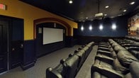 This private home theater is designed to replicate the full cinematic experience with meticulous attention to detail. The room is anchored by a large projection screen framed within a dramatic arched alcove, finished in bold yellow and red tones that contrast with the deep navy blue walls throughout the rest of the space. A ceiling-mounted projector delivers the on-screen presentation, supported by recessed lighting and wall-mounted sconces that provide subtle, atmospheric illumination.

Seating is arranged in tiered rows, elevated by carpeted steps that ensure clear sightlines from every position. Multiple rows of oversized black leather power recliner chairs, each equipped with built-in cup holders, provide a premium viewing experience for a generous number of guests. A designated wheelchair-accessible space is also included at the end of one row. The lower walls feature dark wood wainscoting panels, adding a refined, classic theater aesthetic. Framed vintage movie theater artwork lines the side walls, reinforcing the cinematic theme throughout. Carpeted flooring completes the acoustically considerate design of this well-appointed screening room.