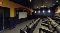 This private home theater is designed to replicate the full cinematic experience with meticulous attention to detail. The room is anchored by a large projection screen framed within a dramatic arched alcove, finished in bold yellow and red tones that contrast with the deep navy blue walls throughout the rest of the space. A ceiling-mounted projector delivers the on-screen presentation, supported by recessed lighting and wall-mounted sconces that provide subtle, atmospheric illumination.

Seating is arranged in tiered rows, elevated by carpeted steps that ensure clear sightlines from every position. Multiple rows of oversized black leather power recliner chairs, each equipped with built-in cup holders, provide a premium viewing experience for a generous number of guests. A designated wheelchair-accessible space is also included at the end of one row. The lower walls feature dark wood wainscoting panels, adding a refined, classic theater aesthetic. Framed vintage movie theater artwork lines the side walls, reinforcing the cinematic theme throughout. Carpeted flooring completes the acoustically considerate design of this well-appointed screening room.