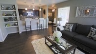 This single-floor residence showcases an open-concept layout that unifies the living, dining, and kitchen areas into one cohesive, flowing space. Rich dark hardwood flooring runs throughout, anchoring the interior with warmth and continuity. The living area is centered around a dark upholstered sofa accented with patterned throw pillows, a glass-top coffee table with decorative metalwork, and a plush cream area rug that adds textural contrast. Natural light filters in through a large window fitted with white blinds, brightening the space considerably. The dining area sits just beyond, illuminated by a ceiling fan with integrated lighting. The kitchen features dark cabinetry paired with a stone-tile backsplash, stainless steel appliances, and a raised breakfast bar with white upholstered bar stools. Pendant lighting above the bar and a track lighting fixture add layered illumination throughout. Neutral gray-toned walls tie each zone together, while the open sightlines between areas create an inviting, spacious atmosphere well-suited for both everyday living and entertaining.