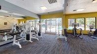 This fitness center at 1160 N Conwell Ave in Covina, California, is a well-appointed, single-floor gym designed to support a comprehensive workout experience. The space is divided into distinct exercise zones, seamlessly connected to create a functional and energizing environment.

The strength training area is outfitted with a variety of cable and weight machines, all finished in white, and is lined with floor-to-ceiling mirrors that amplify the sense of space and allow users to monitor their form. Vibrant yellow accent walls add a bold, motivating pop of color throughout, contrasting with the cool gray and blue tones of the surrounding walls.

The cardio zone features treadmills, elliptical trainers, and stair climbers positioned near large windows and glass doors. These openings provide an abundance of natural light and offer direct views of an outdoor pool area and patio, creating a bright and refreshing atmosphere during workouts. A decorative geometric-patterned accent wall near the entry door adds a contemporary design touch.

Underfoot, durable multi-toned carpet tiles cover the entire floor, providing comfort and traction. Ceiling fans assist with ventilation, and recessed skylights along with flush-mounted light panels ensure consistent illumination. The overall ambiance is modern, clean, and purpose-built to encourage an active lifestyle.