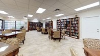 This single-floor community library and activity room offers a welcoming, well-organized environment designed for both leisure and social engagement. The space is generously proportioned, accommodating multiple seating arrangements without feeling crowded. Light-colored large-format tile flooring runs throughout, contributing to a clean, bright aesthetic that complements the neutral walls and drop-panel ceiling fitted with recessed fluorescent lighting panels that evenly illuminate the entire room.

Lining the walls are several tall, dark wood bookshelves filled with a diverse and extensive collection of books, spanning multiple genres and titles. A dedicated display rack near one corner holds a colorful assortment of magazines and periodicals, offering a variety of reading material for all interests.

Throughout the space, multiple dark wood tables are surrounded by elegantly upholstered armchairs featuring a warm, scrolling fabric pattern. These groupings create intimate areas suited for reading, board games, or casual conversation. At least one table is set up for chess or checkers, highlighting the room's recreational purpose.

Windows fitted with horizontal blinds allow natural light to filter in, and framed artwork adorns the walls, adding character and warmth to this inviting, community-oriented gathering space.