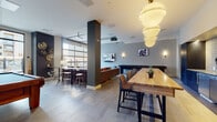 This contemporary club room is thoughtfully designed to serve as a multifunctional social hub, seamlessly blending entertainment, relaxation, and casual dining into one cohesive space. The open-concept layout is anchored by large industrial-style garage windows and full-height glass doors that flood the interior with natural light and offer views of the surrounding area.

The space is divided into distinct yet connected zones. A game area houses a full-size pool table and a dedicated shuffleboard table, both positioned to allow comfortable play. Adjacent to these is a relaxed lounge area furnished with plush sectional sofas, accent chairs, and a sculptural coffee table, centered around a wall-mounted flat-screen TV.

A bar-height communal table with leather stools provides an ideal gathering spot, complemented by a well-equipped kitchenette featuring dark navy cabinetry, a patterned tile backsplash, and pendant lighting. A separate high-top bar area with barstool seating adds another social node to the room.

Throughout, the space features large-format tile flooring, dark accent columns, layered wall sconce lighting, and distinctive pendant fixtures that enhance the modern-industrial aesthetic. Decorative wall art and carefully chosen furnishings complete the refined yet approachable ambiance, making this club room a versatile venue for both lively gatherings and quieter social moments.