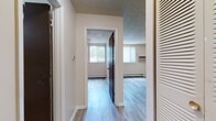 This single-floor unit offers a straightforward and functional layout designed for comfortable everyday living. Light-toned hardwood-style flooring runs consistently throughout the space, creating a cohesive and easy-to-maintain interior. A central hallway connects the main areas, with louvered closet doors providing convenient storage access near the entry.

The kitchen is equipped with warm honey-toned wood cabinetry, a white electric coil range with overhead ventilation, and a full-size refrigerator. A sink with countertop workspace completes the kitchen, making it practical for daily meal preparation. The open sightline between the kitchen and the adjoining living area adds a sense of openness to the space.

The living area is generously proportioned, with neutral-toned walls and a large window fitted with vertical blinds that allow ample natural light to filter in. A baseboard heating unit runs along the perimeter, ensuring consistent warmth. A wall-mounted air conditioning unit provides cooling during warmer months.

The bedroom is a comfortable, well-lit room featuring its own window for natural light and outdoor views, a ceiling fan, and baseboard heating. The textured popcorn ceilings are present throughout the unit, adding a classic character consistent with the overall aesthetic.

The bathroom includes a full-size soaking tub enclosed with clean white square tile surround and chrome fixtures, offering a simple and functional bathing space.