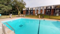 This single-story community facility offers a well-maintained outdoor pool area and updated restroom facilities. The outdoor space centers around a generously sized swimming pool with clear turquoise water, accessible via built-in entry rails. The pool deck is equipped with lounge chairs and a patio umbrella, providing comfortable seating options for residents. A secure chain-link fence encloses the pool area, ensuring safety and privacy. The surrounding landscape features mature trees, offering natural shade, while a multi-story brick residential building serves as the backdrop, its balconies overlooking the pool area.

The interior restroom facilities are finished to a high standard, featuring bold black subway tile wainscoting that contrasts sharply with crisp white upper walls and light gray tile flooring. Stainless steel fixtures throughout — including partition panels, paper towel dispensers, and waste receptacles — lend a clean, durable aesthetic. The vanity area includes a white porcelain sink with a chrome faucet, a silver-framed mirror, and stainless steel accessories. Private stall areas are enclosed with polished metal partitions, and a solid wood-grain door with a chrome lever handle provides secure room access.