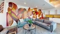 Located at 622–626 Third Ave S in Nashville, TN, this single-floor living room lounge is a thoughtfully designed communal space that blends artistic flair with modern functionality. The room's most striking feature is a sweeping, large-scale abstract mural in warm tones of red, orange, gold, and burgundy, commanding an entire wall and setting a bold, energetic atmosphere throughout the space.

Polished concrete floors run throughout, lending an industrial yet refined character that complements the clean white walls and exposed concrete ceiling details. The lounge area is anchored by a pair of contemporary gray sofas arranged around a round glass-top coffee table, creating a comfortable and inviting gathering spot. A wall-mounted flat-screen TV and a wall-unit HVAC system add practical amenities, while a sleek leather accent chair rounds out the seating.

A built-in kitchenette lines one wall, featuring white cabinetry, open shelving, a dark countertop, a black matte sink, warm yellow tile backsplash, and a stainless steel beverage refrigerator — ideal for entertaining or communal use.

Floor-to-ceiling glass sliding doors flood the space with natural light and provide direct access to a shared outdoor deck, offering views of the surrounding Nashville architecture and seamlessly connecting the interior to the exterior.
