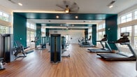 This single-floor fitness center is designed to support a comprehensive workout experience, combining thoughtful aesthetics with a full range of exercise equipment. The space is defined by striking teal accent walls paired with crisp white trim, creating a bold and energizing atmosphere. Warm hardwood-style flooring runs throughout, providing both visual warmth and a durable surface suited for a variety of training activities.

Large, multi-pane windows line the walls, flooding the interior with abundant natural light and offering views of the surrounding outdoor environment. The bright, open feel is further enhanced by recessed ceiling lighting and ceiling fans that maintain comfortable airflow during workouts.

The cardio section is well-appointed with multiple treadmills and elliptical machines, each equipped with individual display screens. The strength training area features a selection of selectorized weight machines, cable systems, and benches, providing options for both upper and lower body conditioning. A large wall-mounted flat-screen television is positioned for easy viewing from the strength training zone.

Full-length mirrors are strategically placed to visually expand the space and assist users in monitoring their form. The layout is open and uncluttered, allowing for ease of movement between equipment stations. The overall design balances functionality with a clean, modern aesthetic, making this fitness center an inviting and practical space for daily exercise.